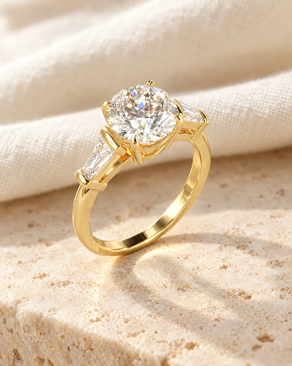 Verlobungsring Valerie: runder Diamant zwischen zwei Tapered-Baguette-Diamanten, Gelbgold, auf hellem Travertin